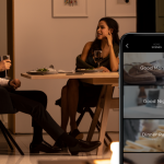 A couple has dinner in a warmly illuminated room while a phone shows different scenes in the Savant app.