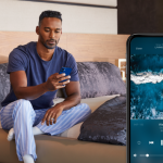 Man sitting on a modern bed, using a smartphone to control music playback, with a close-up of the phone screen showing a streaming app playing ocean sounds.